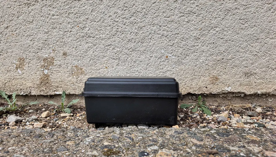 Boîtier d'appâtage noir fermé posé au pied d'un mur crépi, sur du gravier